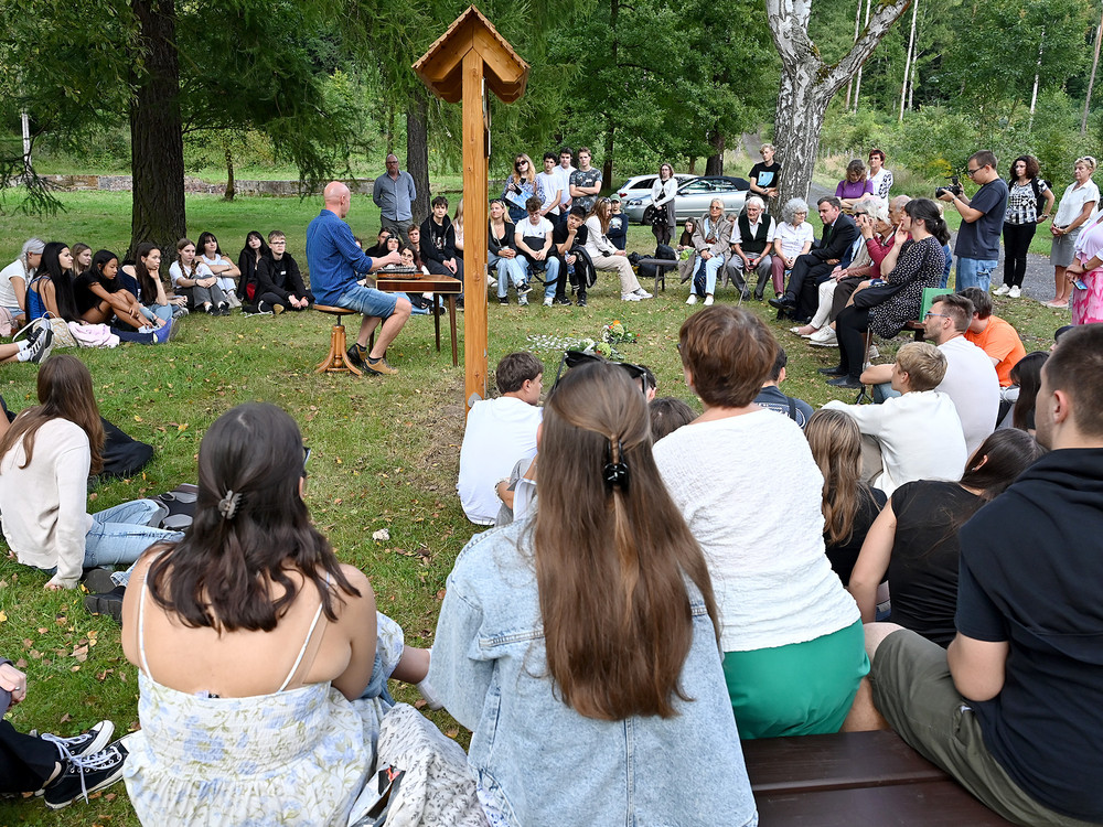 KI generiert: Das Bild zeigt eine Gruppe von Personen im Freien, wobei zwei junge Männer im Vordergrund stehen und sich unterhalten, während eine Informations-tafel und Wegweiser im Hintergrund zu sehen sind. Der Hauptinhalt des Bildes scheint eine Diskussion oder Führung in einer natürlichen Umgebung, möglicherweise einem Park oder Wald, zu sein.
