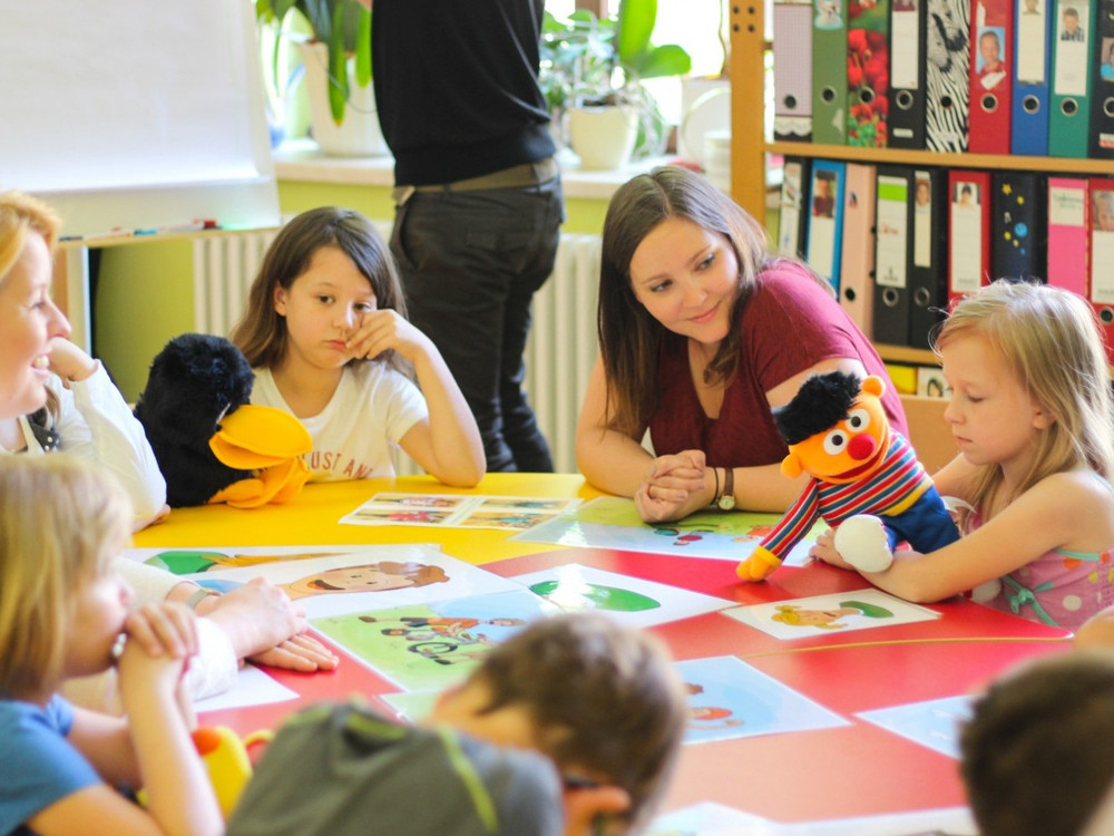 KI generiert: Das Bild zeigt eine Gruppe von Kindern und Erwachsenen, die an einem Tisch sitzen und mit Spielzeug und Bildern spielen oder arbeiten. Der Hauptinhalt des Bildes ist eine interaktive und pädagogische Aktivität, möglicherweise in einem Kindergarten oder einer Vorschule.