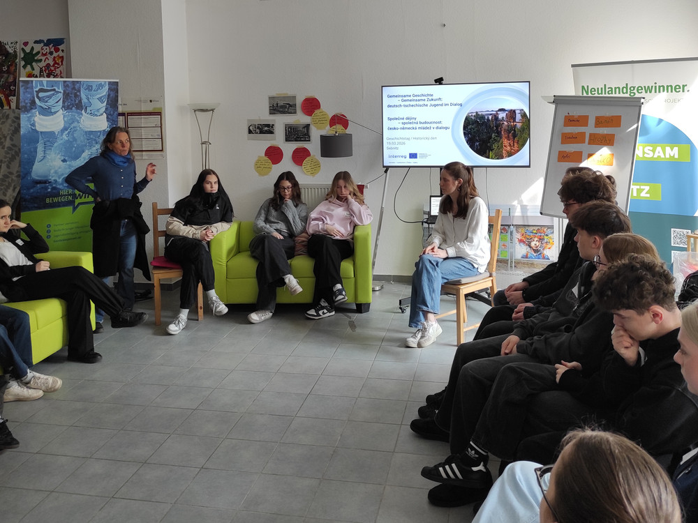KI generiert: Das Bild zeigt eine Gruppe von Personen in einem Klassenzimmer, wobei eine Person vor einem Whiteboard steht und eine Präsentation hält. Der Hauptinhalt des Bildes ist eine Unterrichts- oder Besprechungssituation, in der eine Präsentation oder Diskussion stattfindet.
