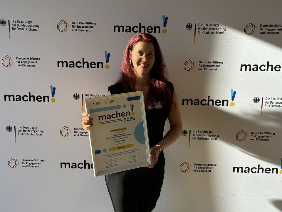 KI generiert: Das Bild zeigt eine Person, die vor einem Step-and-Repeat-Banner mit dem Aufdruck "machen!" und verschiedenen Logos steht und ein Zertifikat oder eine Urkunde mit der Aufschrift "machen!2026" in der Hand hält. Der Hauptinhalt des Bildes ist die Präsentation oder Auszeichnung, die die Person erhalten hat.