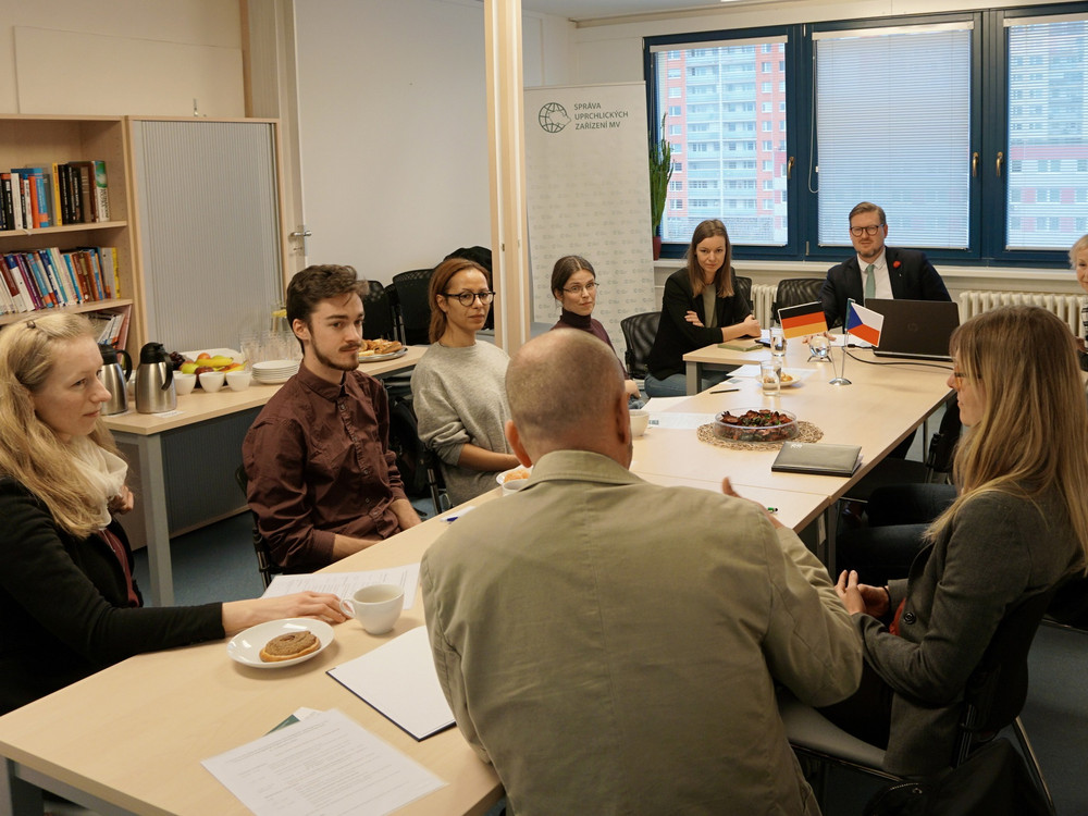KI generiert: Das Bild zeigt eine Gruppe von Personen, die in einem Besprechungsraum an einem langen Tisch sitzen und sich unterhalten. Im Vordergrund sind einige Teilnehmer im Gespräch, während im Hintergrund weitere Personen am Tisch sitzen, darunter eine Person mit kleinen Flaggen vor sich.