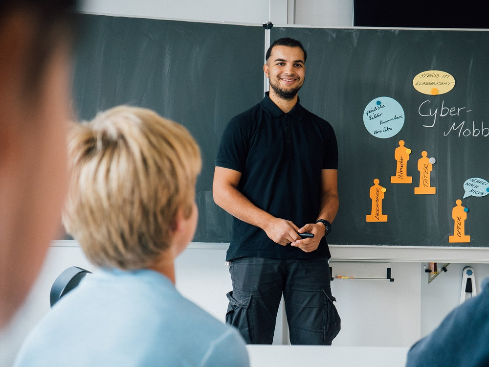 KI generiert: Das Bild zeigt einen Mann, der vor einer Tafel steht und einer Gruppe von Personen eine Präsentation über Cyber-Mobbing hält. Die Tafel enthält verschiedene Notizen und Zeichnungen, die sich auf das Thema beziehen.