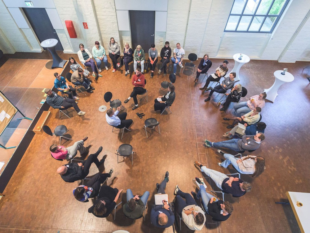 KI generiert: Das Bild zeigt eine Gruppe von Menschen, die in einem großen, hellen Raum mit Holzboden in einem Kreis sitzen und sich unterhalten. Im Vordergrund sind einige Personen, die auf Stühlen sitzen, während andere auf dem Boden Platz genommen haben, was auf eine informelle Diskussion oder ein Meeting hindeutet.