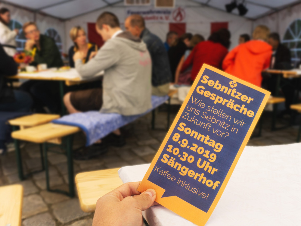 KI generiert: Das Bild zeigt eine Person, die eine Einladungskarte für ein "Sebnitzer Gespräch" hält, das am Sonntag, den 8.9.2019 um 10:30 Uhr im Sängerhof stattfindet, inklusive Kaffee. Im Hintergrund sind Menschen in einem Zelt, die sich unterhalten und an Tischen sitzen.