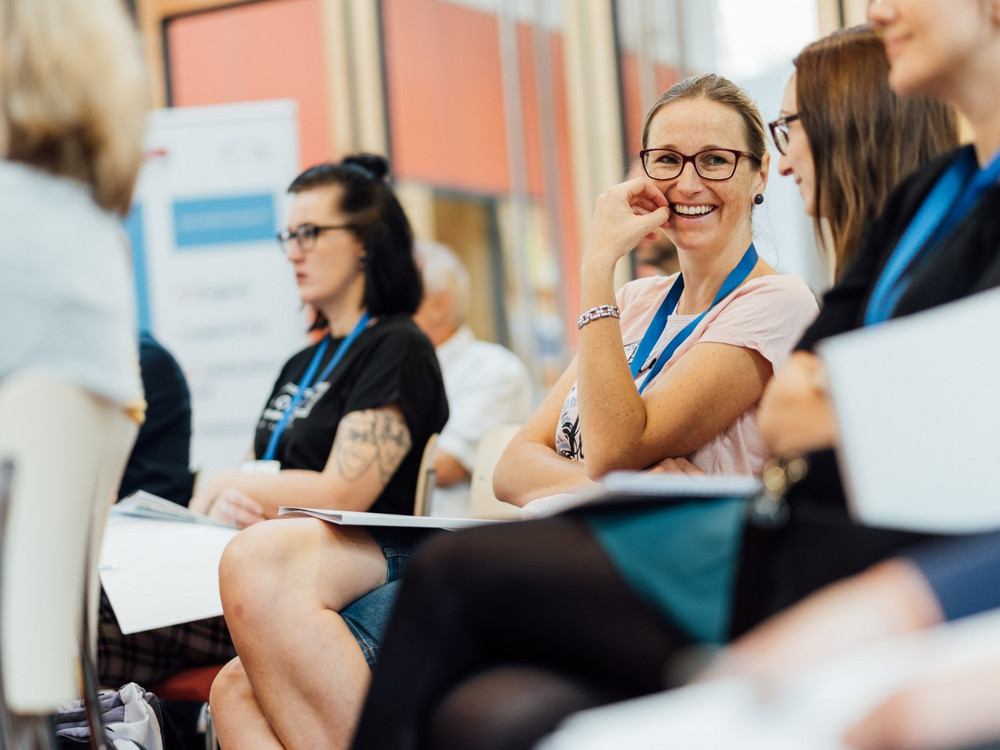 KI generiert: Das Bild zeigt eine Gruppe von Personen, die in einer informellen Sitzung oder einem Workshop zusammenkommen. Die Teilnehmer halten Notizblöcke und tragen Namensschilder, was auf eine organisierte Veranstaltung oder Konferenz hindeutet.