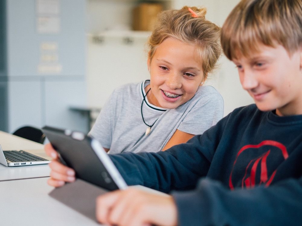 KI generiert: Das Bild zeigt zwei Kinder, die gemeinsam an einem Tisch sitzen und sich auf ein Tablet konzentrieren, während ein Laptop in ihrer Nähe steht. Der Hauptinhalt des Bildes ist die Interaktion der Kinder mit dem Tablet, was auf eine Lern- oder Spielsituation hindeuten könnte.